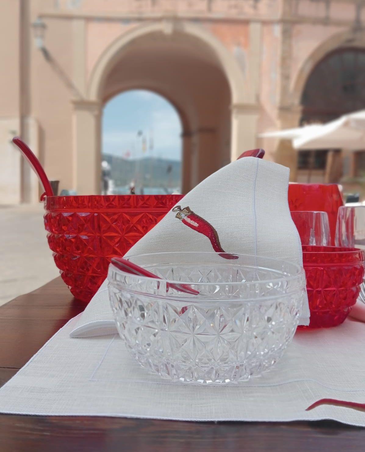 dettaglio elegante del tovagliolo in lino cerato ricamato con corno rosso su tavola estiva, con ciotole acriliche  Mario Luca Giusti e vista sul porto