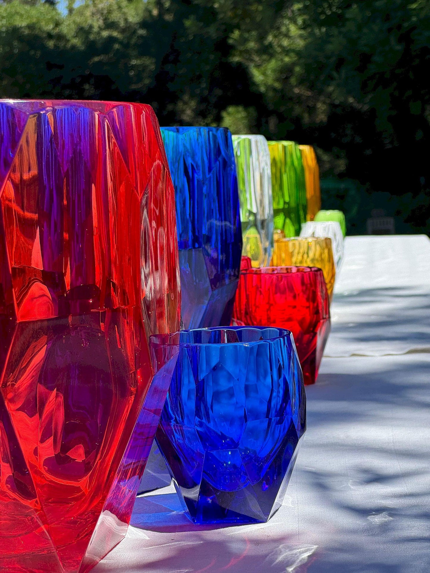 Porta bottiglie Antartica di colori diversi con bicchieri Super Milly di vari colori di Mario Luca Giusti, in cristallo acrilico, fotografati all’aperto su tavolo estivo, design sfaccettato elegante e colorato