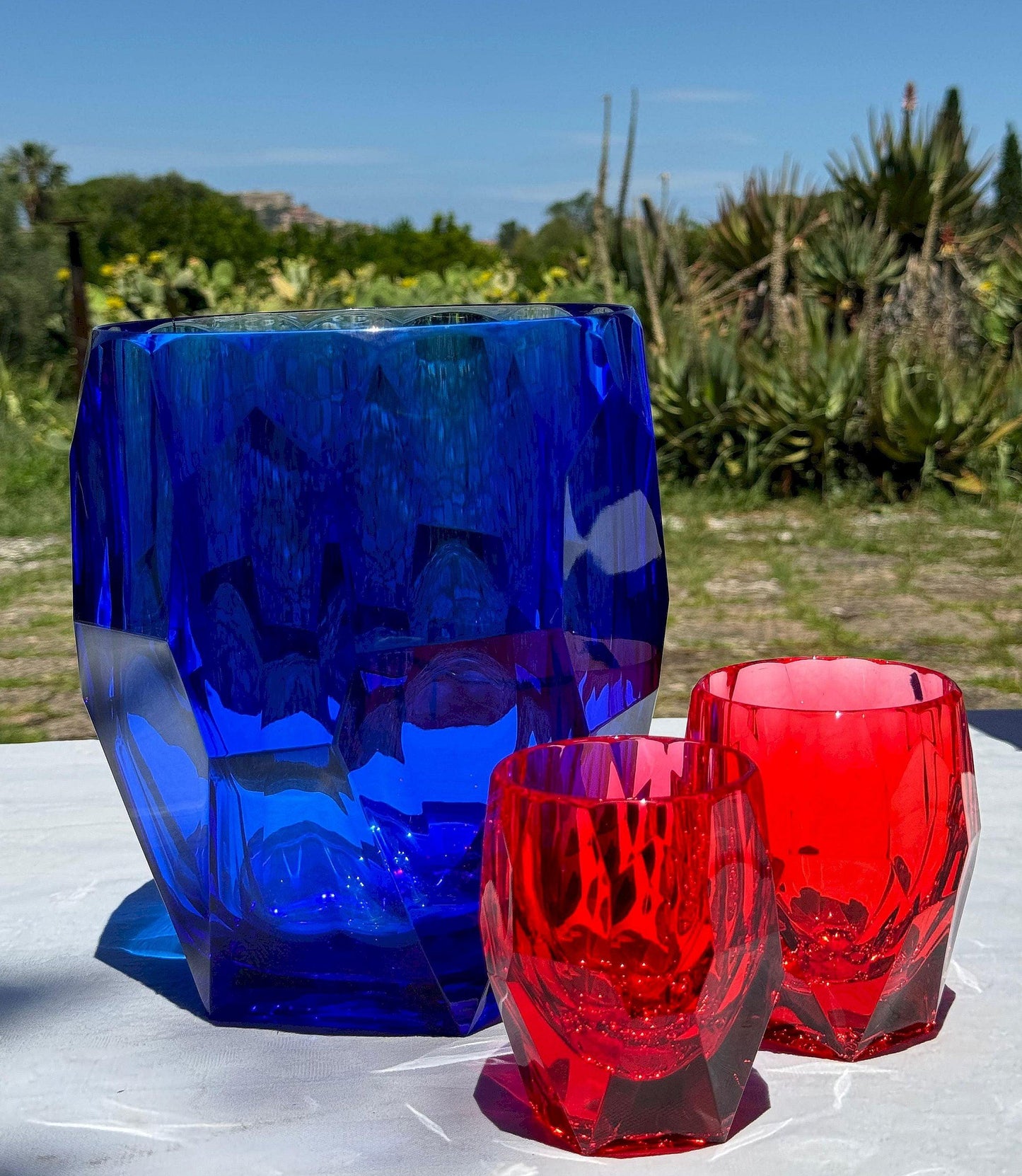 Porta bottiglie Antartica blu con bicchieri Milly e Super Milly rossi di Mario Luca Giusti, in cristallo acrilico, fotografati all’aperto su tavolo estivo, design sfaccettato elegante e colorato