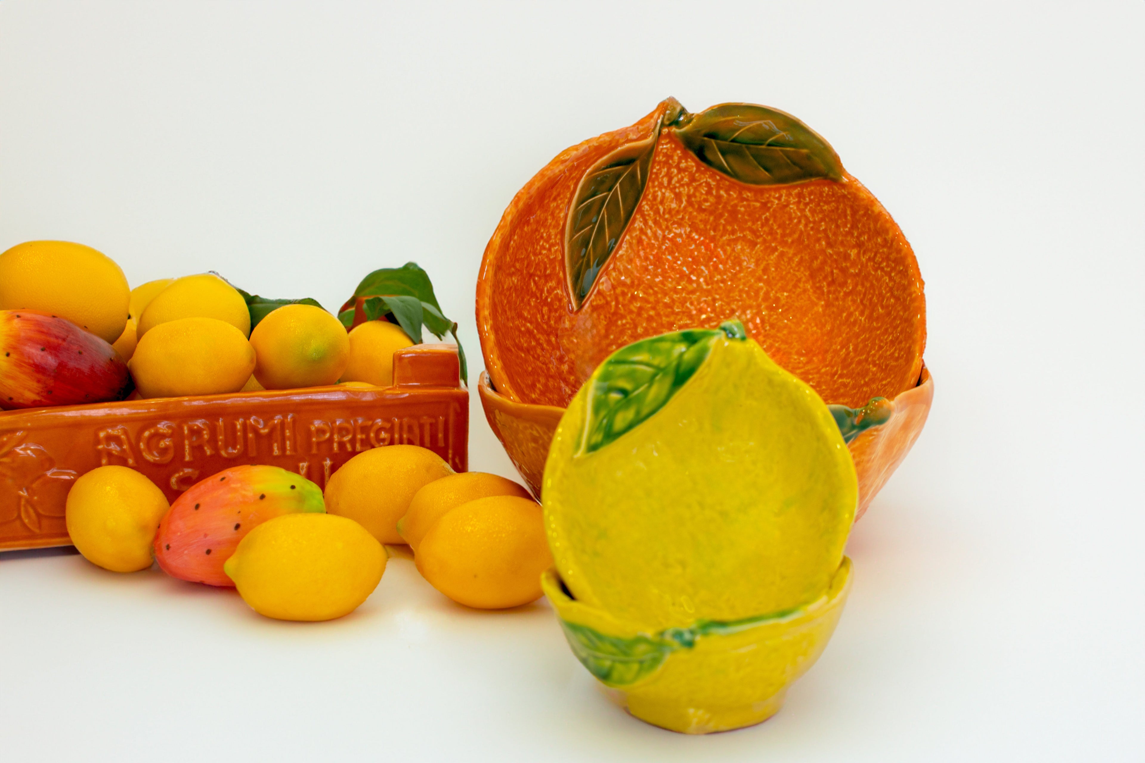 Ciotole in ceramica a forma di limone e arancia con decorazioni a rilievo, accanto a una cassetta di agrumi e fichi d’India decorativi