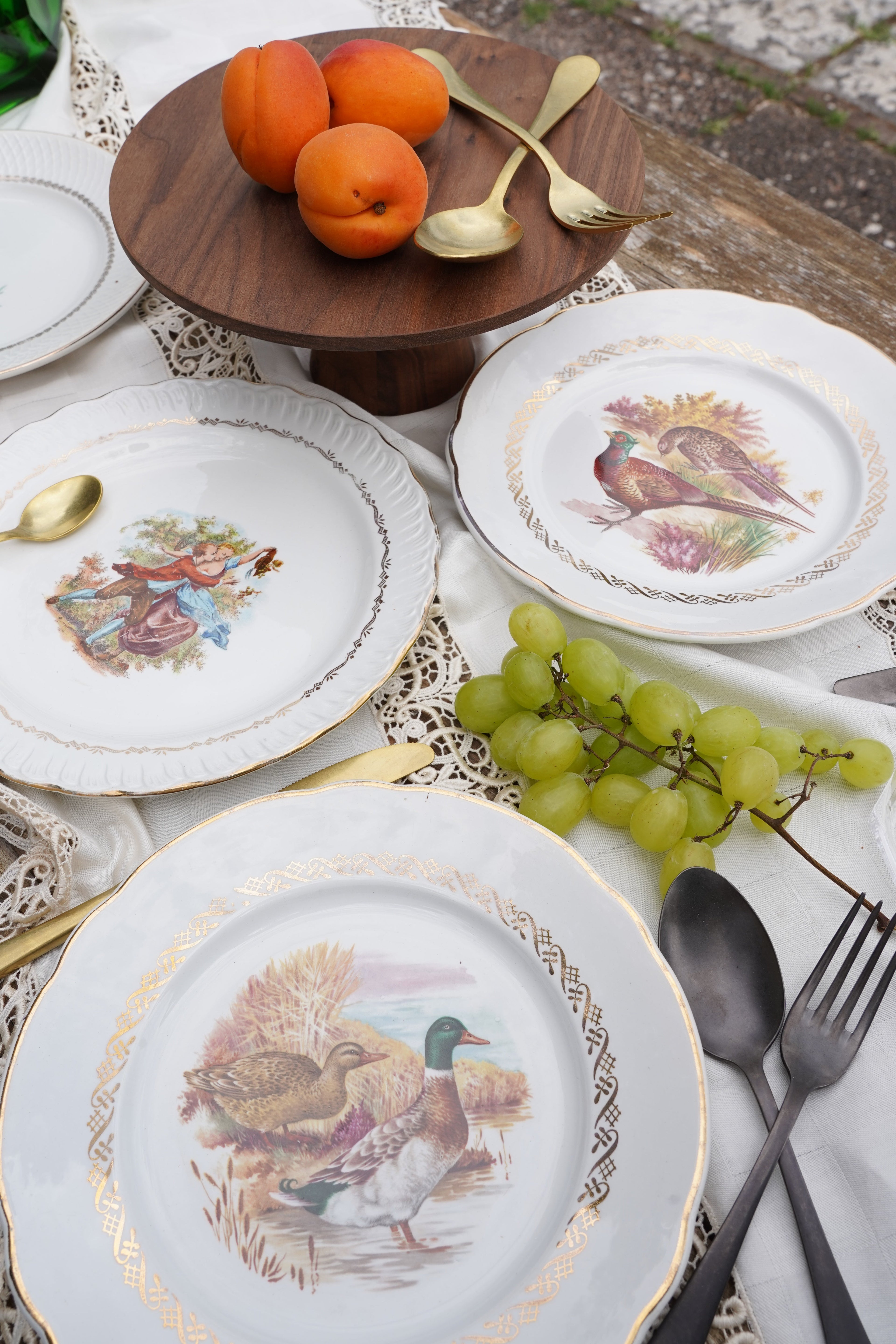 Piatti vintage decorati con scene romantiche e animali, bordo dorato, apparecchiati su tavola elegante con frutta e alzata in legno