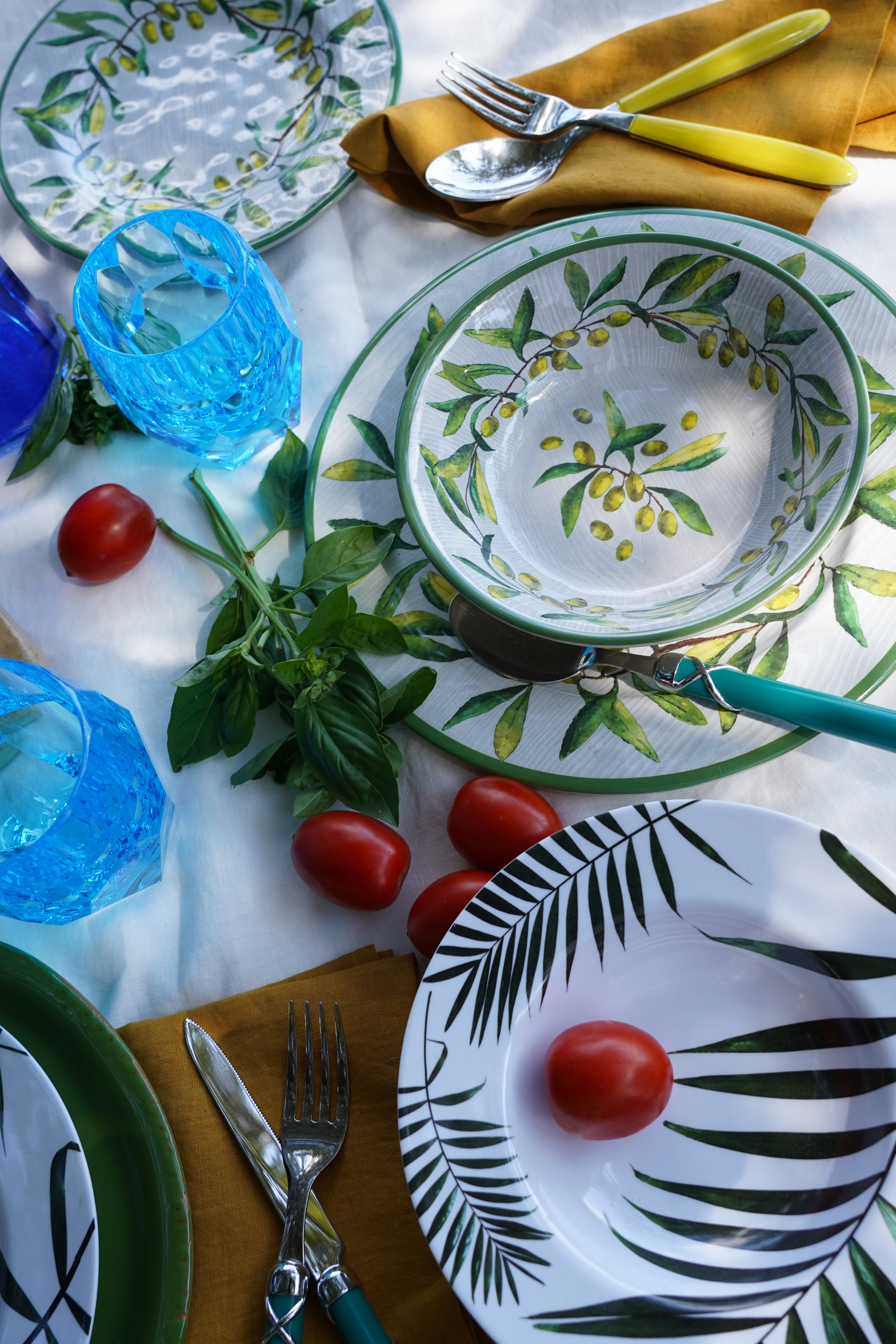 Set di piatti in melamina con decorazioni tropicali e ulivo, apparecchiati su tavola estiva con bicchieri blu, posate e pomodorini