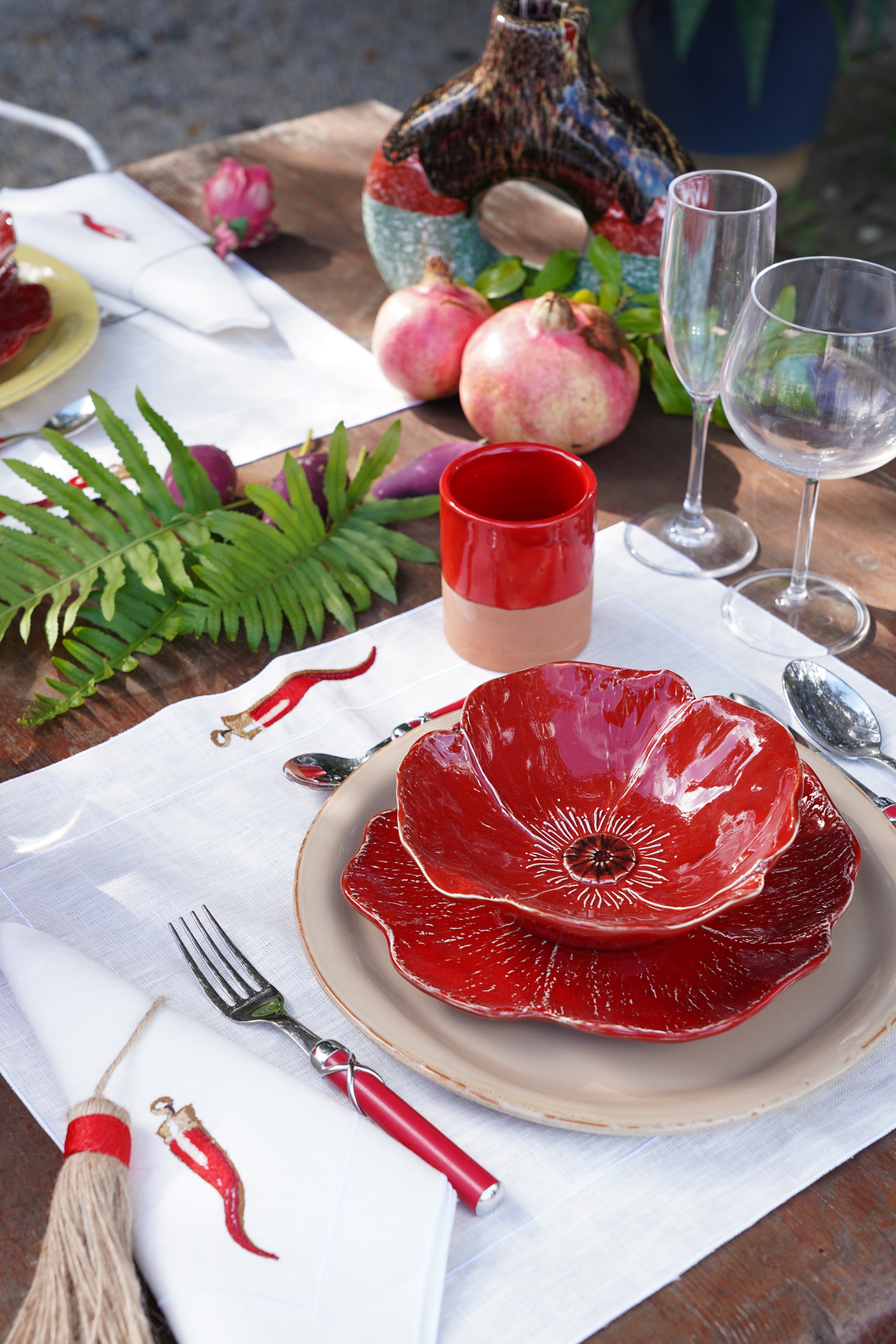 Piatto in ceramica artigianale a forma di fiore rosso su tavola apparecchiata, con sottopiatto neutro, bicchieri coordinati e dettagli naturali