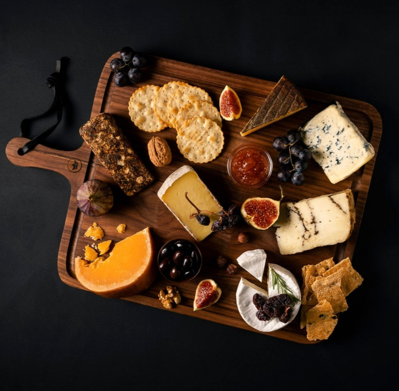 Tagliere in legno con selezione di formaggi assortiti, fichi, uva, cracker, frutta secca e marmellata.