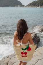 Donna di spalle con borsa in paglia Cherry Marika Selection con ricamo ciliegie rosse e nappa decorativa, indossata su scogli vista mare, stile estivo boho chic.