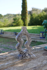 Vaso decorativo artigianale in ceramica, design scultoreo sinuoso bianco e nero, posato su tavolo in legno con sfondo naturale.