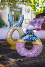 Due vasi decorativi in ceramica smaltata, uno giallo e blu dalla forma biforcuta e uno rosa, giallo e blu dalla forma circolare con foro centrale, fotografati su un tavolo in esterno.