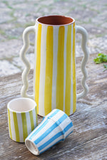 Vaso caraffa in ceramica fatto a mano con righe giallo e bianco, altezza 28 cm, con piccoli bicchieri artigianali su tavolo rustico