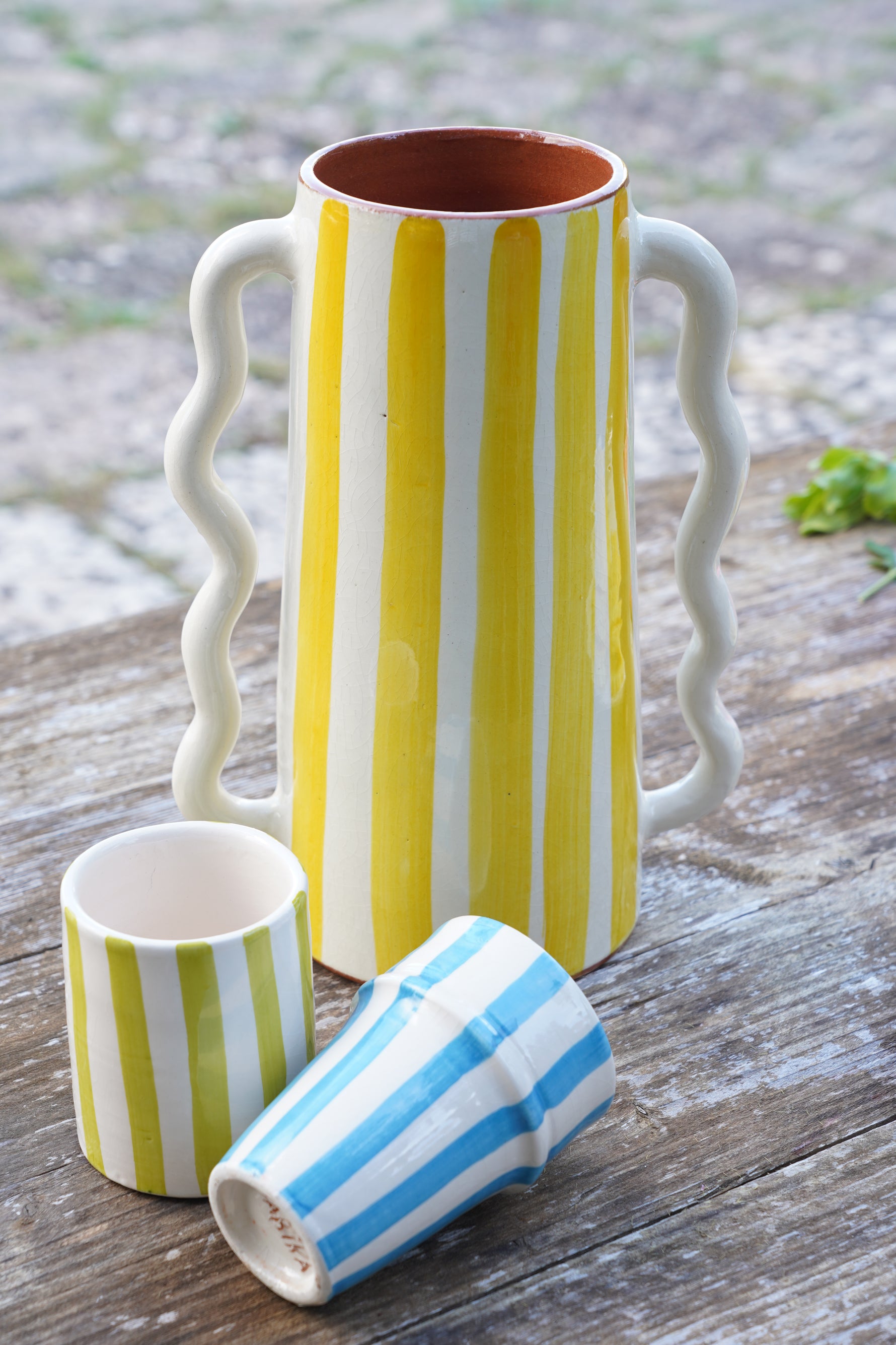 Caraffa in ceramica artigianale a righe gialle con doppio manico ondulato, accompagnata da bicchieri coordinati a righe su tavolo in legno all’aperto