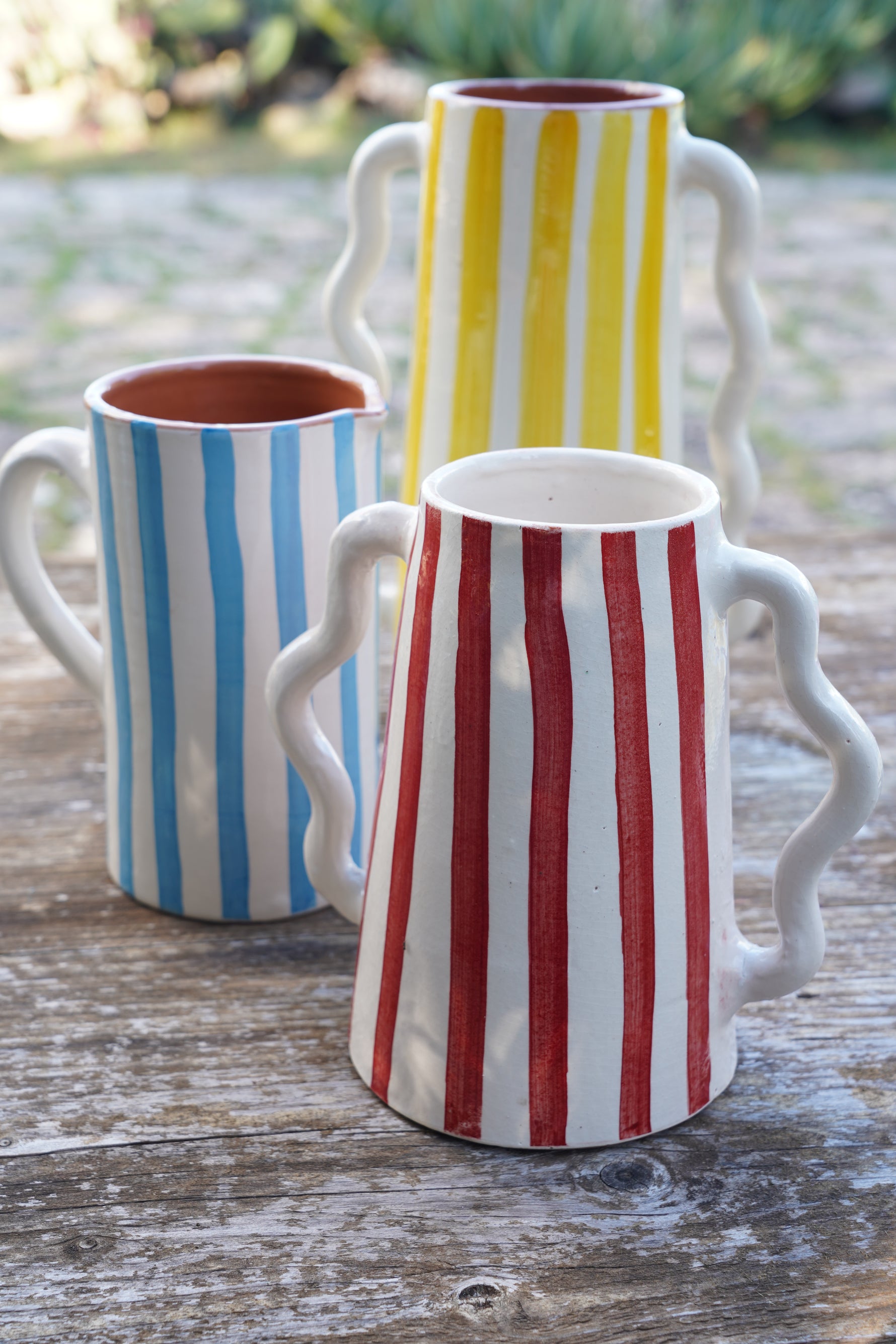 Brocche in ceramica artigianale a righe blu, gialle e rosse su tavolo in legno all’aperto