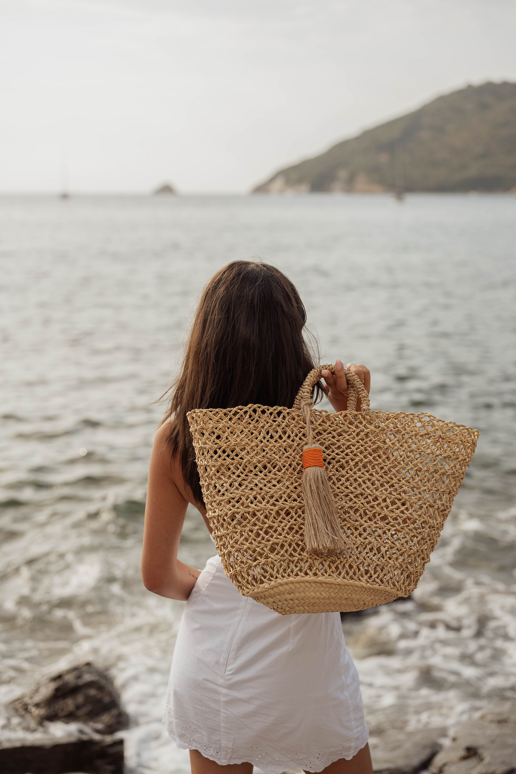 Donna di spalle con borsa in paglia intrecciata Marika Selection con nappa decorativa, vista mare, stile estivo naturale ed elegante.