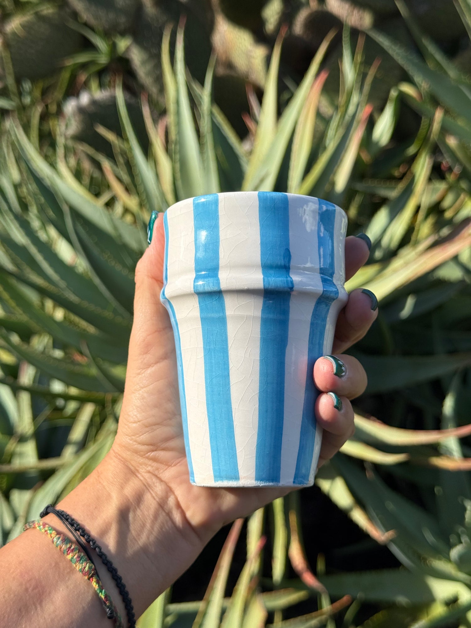 Bicchiere in ceramica artigianale con righe azzurre verticali, tenuto in mano davanti a piante grasse
