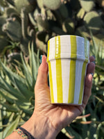 Bicchiere in ceramica artigianale con righe verde chiaro, tenuto in mano davanti a cactus e piante succulente