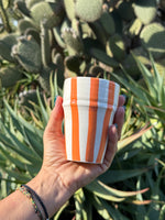 Bicchiere in ceramica artigianale con righe arancioni verticali, tenuto in mano davanti a piante grasse
