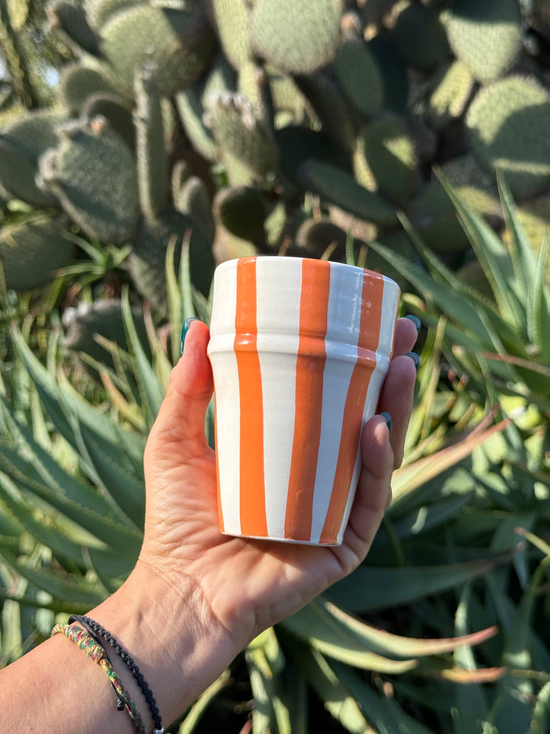 Bicchiere in ceramica artigianale con righe arancioni verticali, tenuto in mano davanti a piante grasse