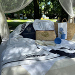teli mare bianchi con bordo e ricamo nodo marino blu, artigianali made in Italy, esposti con accessori estivi e piatti decorativi su letto outdoor