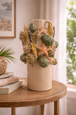 Vaso anfora in ceramica con conchiglie e stelle marine in rilievo, ambientato su tavolo in legno in un interno luminoso stile mediterraneo