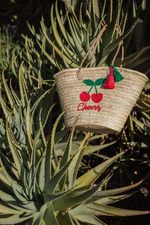 Borsa in paglia Cherry Marika Selection con ricamo ciliegie rosse e nappa decorativa appoggiata su piante di cactus, stile estivo boho chic naturale.