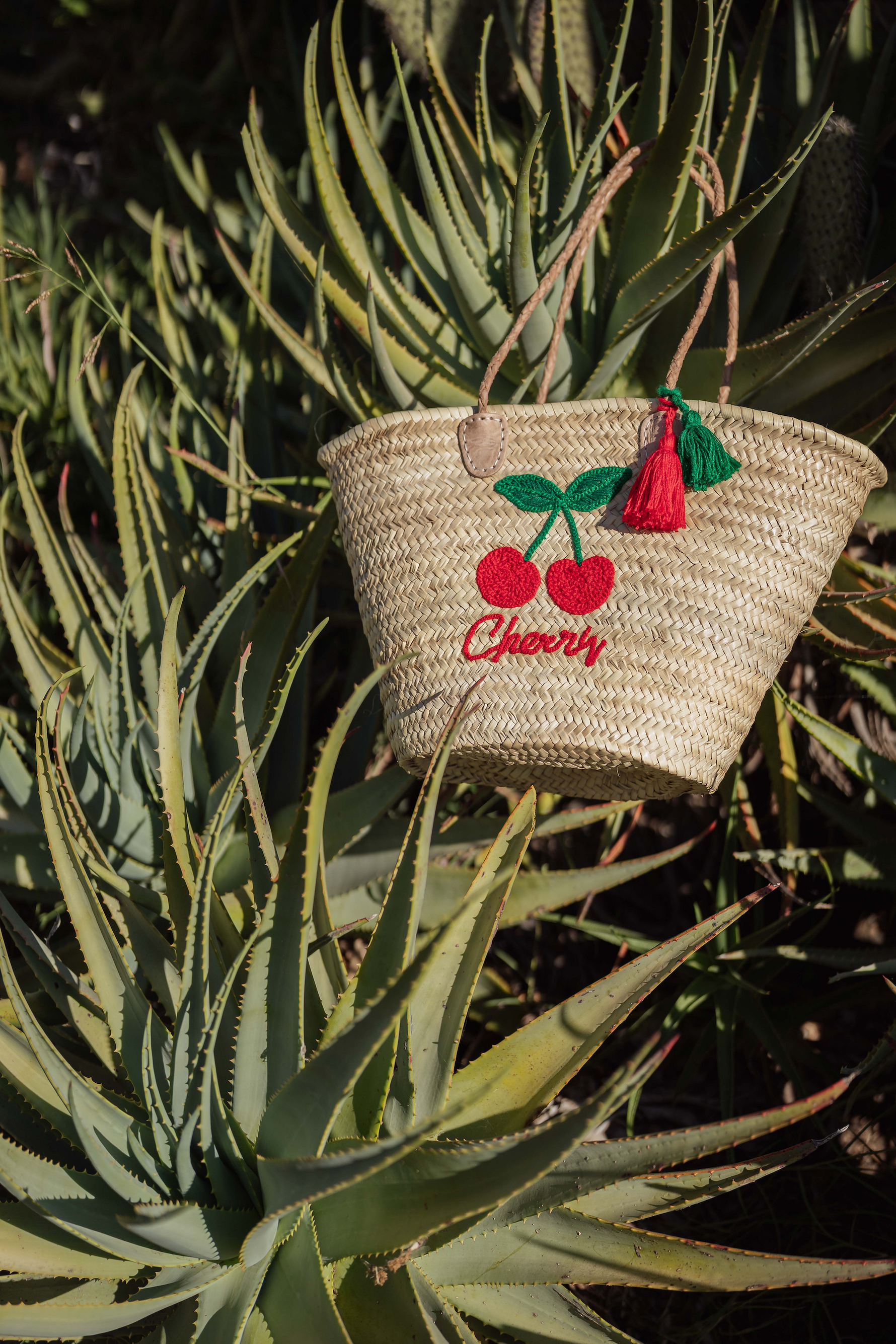 Borsa in paglia Cherry Marika Selection con ricamo ciliegie rosse e nappa decorativa appoggiata su piante di cactus, stile estivo boho chic naturale.