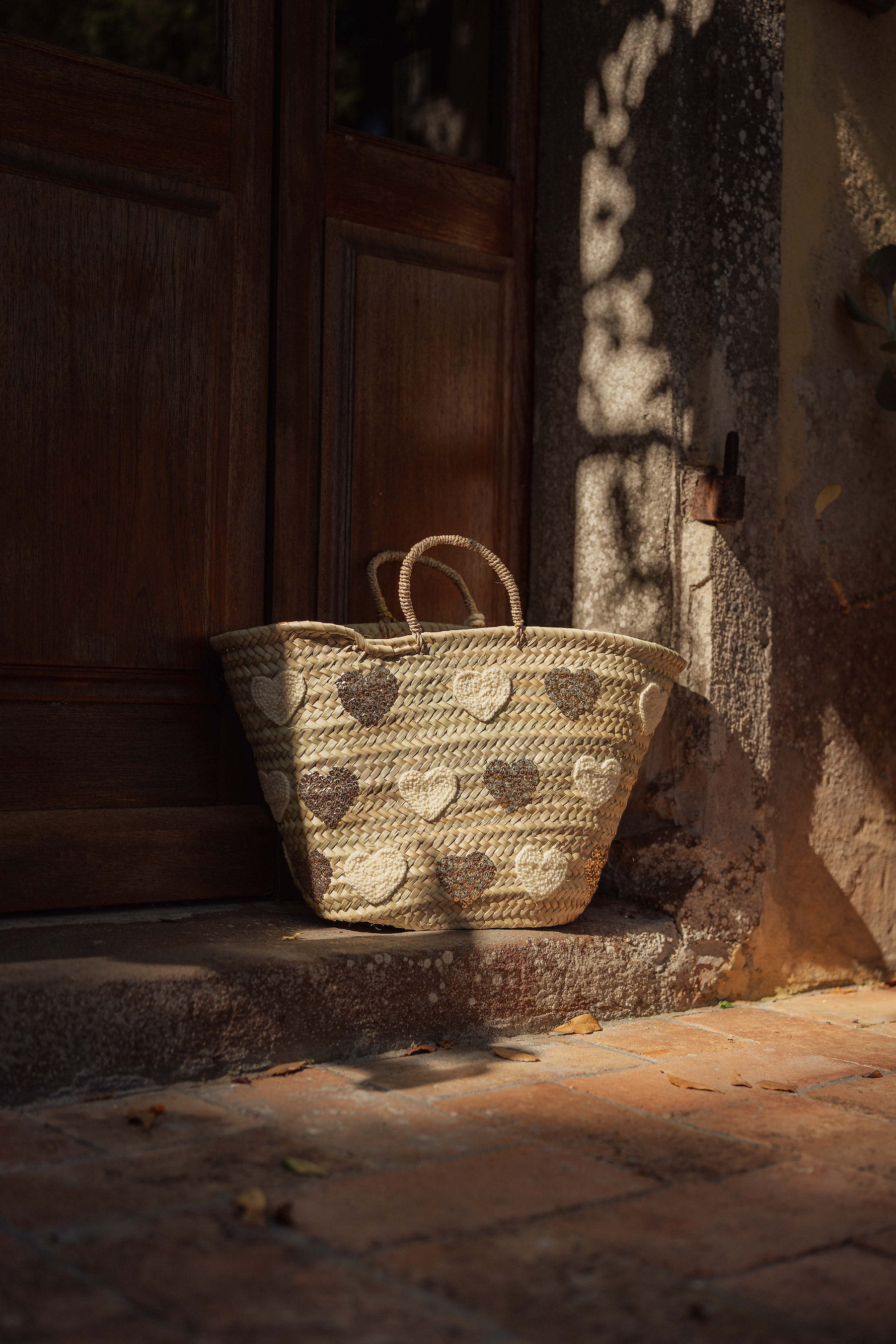 Borsa in paglia intrecciata con cuori ricamati naturali e dorati appoggiata a porta, Marika Selection