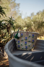 Borsa in paglia ricamata con fiori viola e bianchi Marika Selection appoggiata su fontana in giardino, stile estivo boho chic naturale ed elegante.