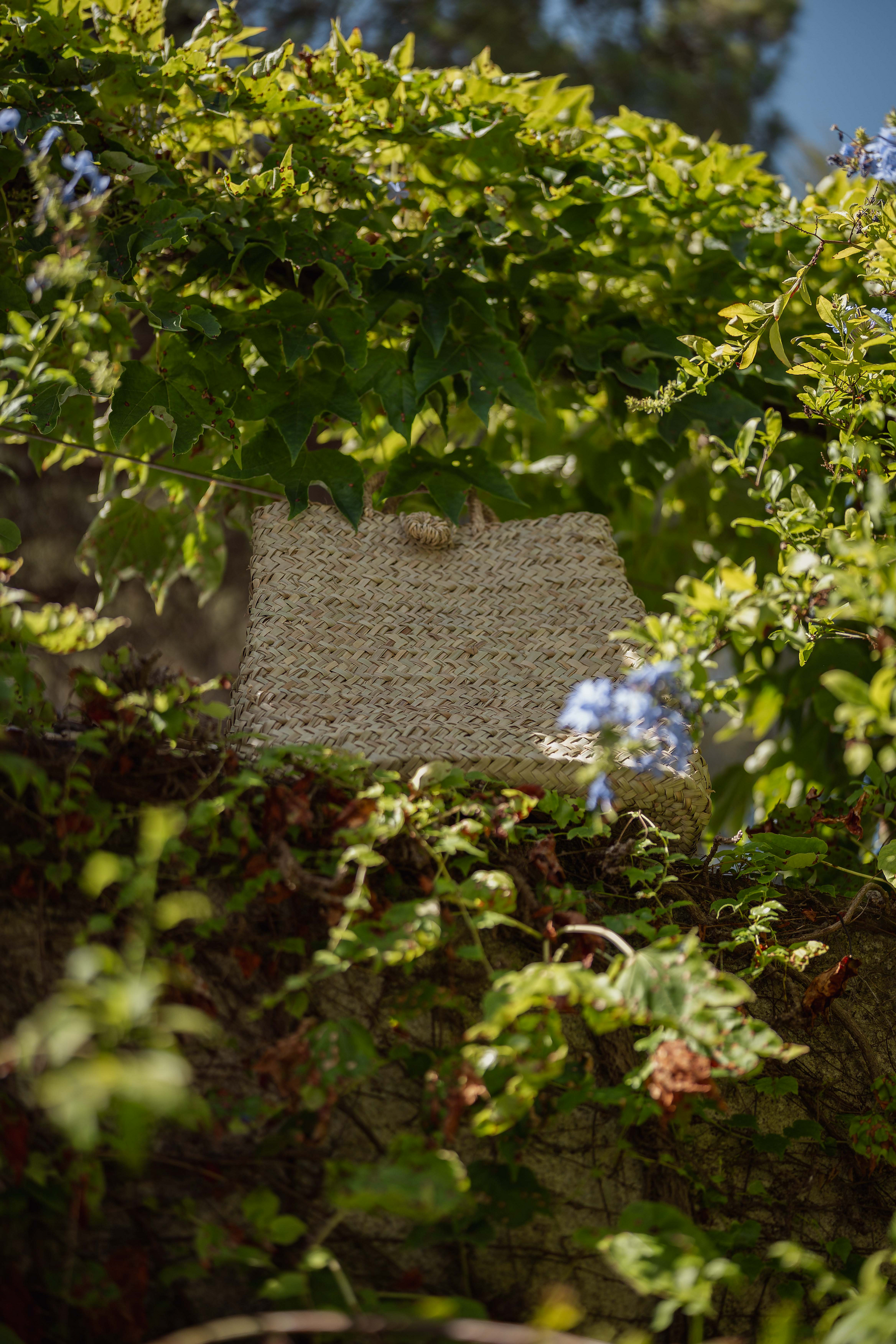 Borsa in paglia naturale Marika Selection appoggiata tra piante verdi in giardino, accessorio estivo elegante e naturale.

