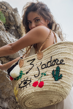 Donna con borsa in paglia J’Adore ricamata con scritta, ciliegie, cactus e doppia nappa panna e nera, vista mare su scogli.