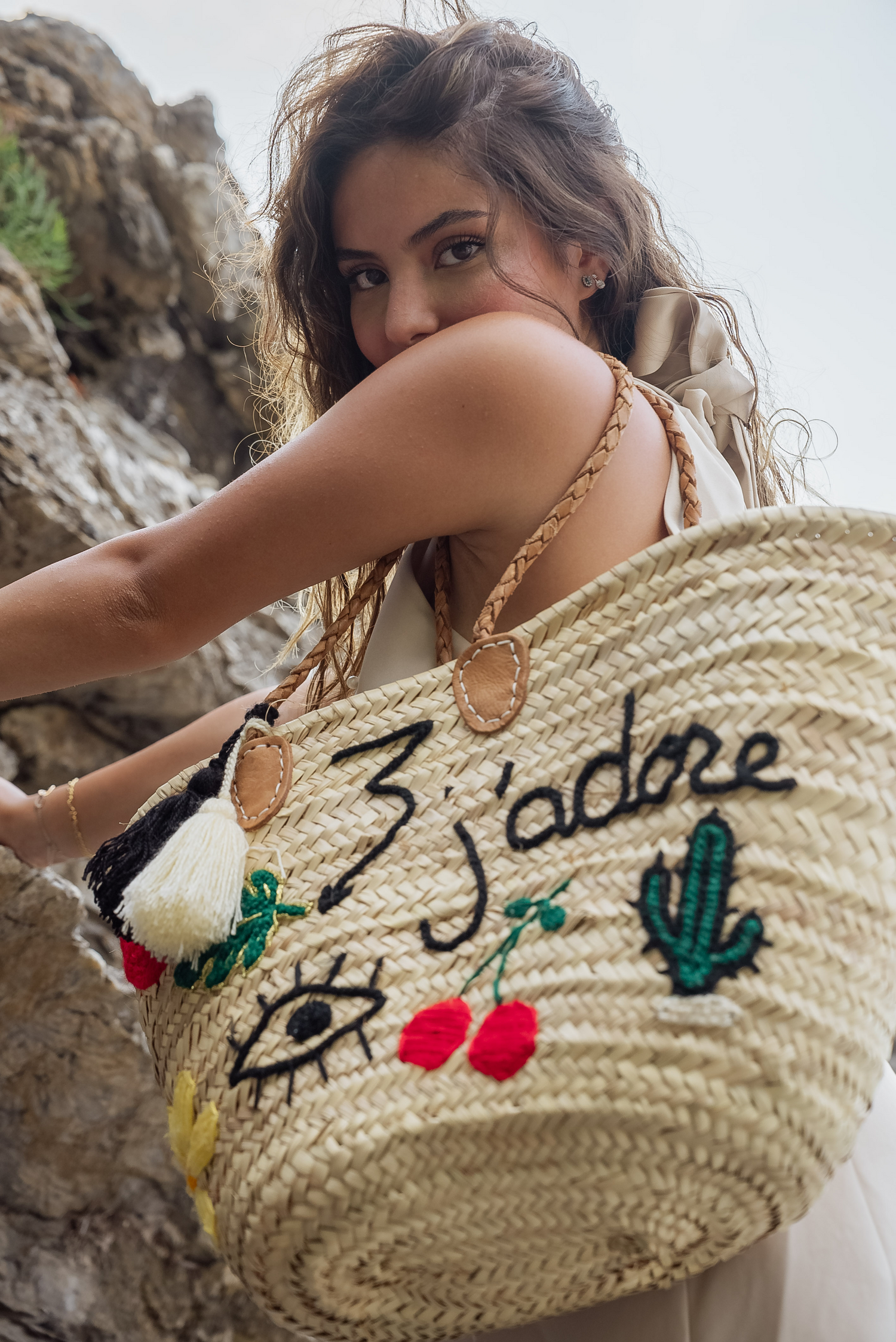 Donna con borsa in paglia J’Adore ricamata con scritta, ciliegie, cactus e doppia nappa panna e nera, vista mare su scogli.
