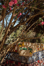 Borsa in paglia “Dolce Vita” con limoni ricamati e nappe bicolore appesa a rami in giardino mediterraneo.