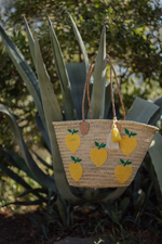 Borsa in paglia Marika Selection con ricamo limoni gialli e doppia nappa decorativa gialla e bianca appoggiata tra piante di cactus, stile estivo.

