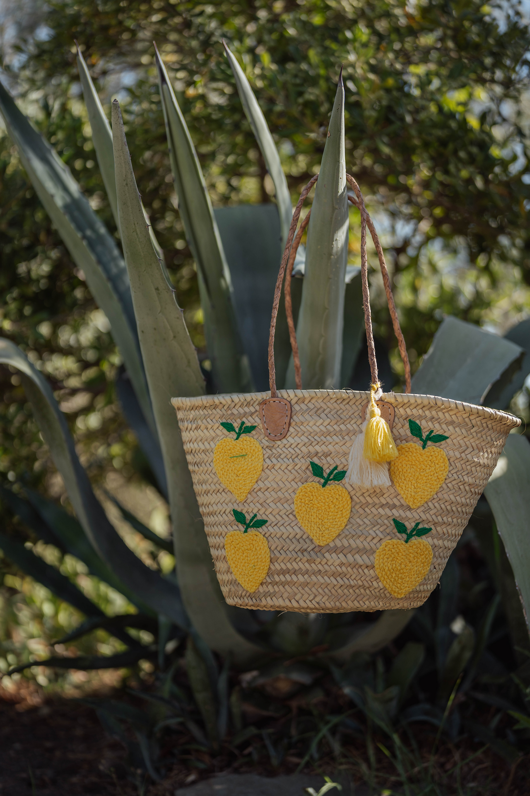 Borsa in paglia Marika Selection con ricamo limoni gialli e doppia nappa decorativa gialla e bianca appoggiata tra piante di cactus, stile estivo.
