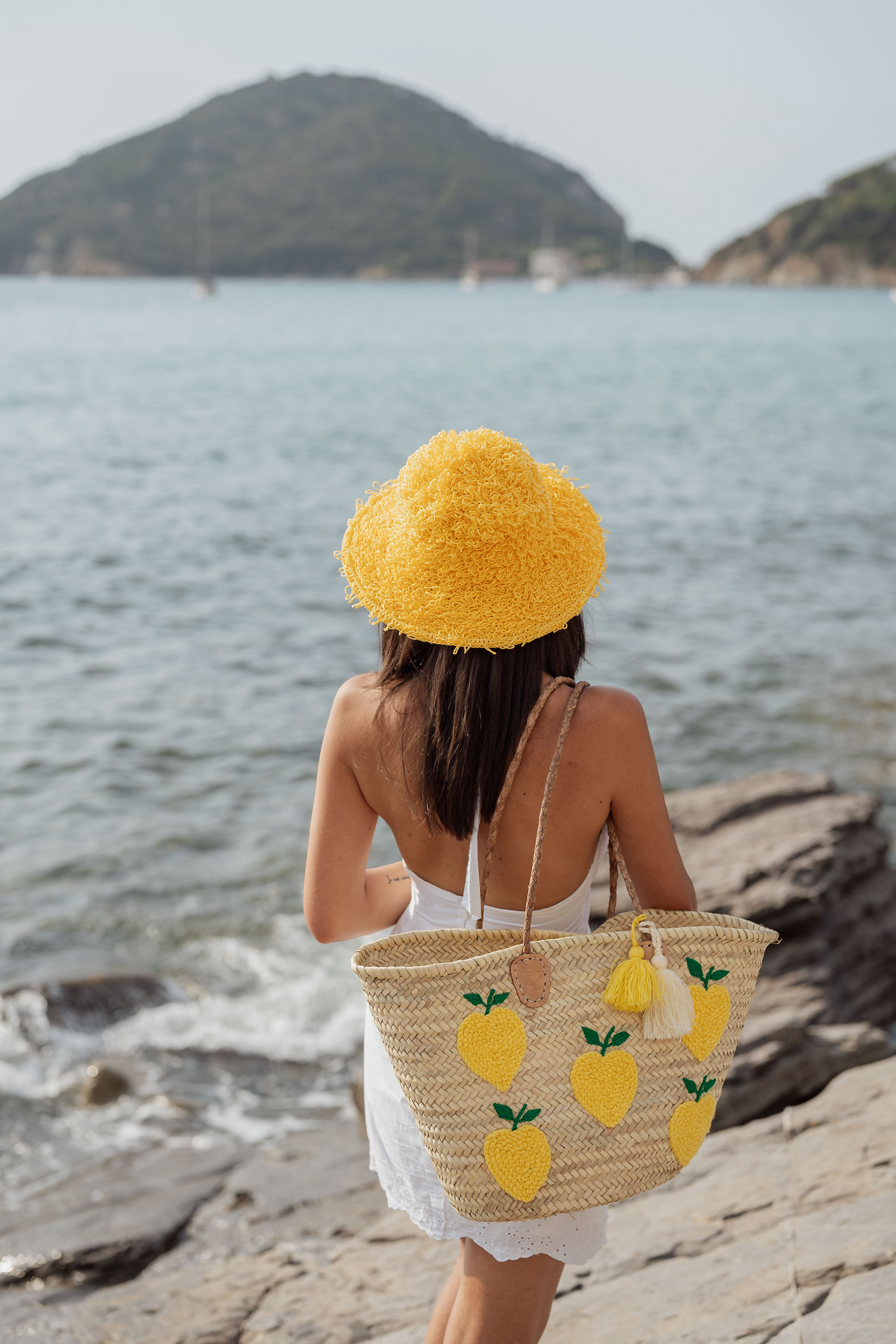 Donna di spalle con borsa in paglia Marika Selection con ricamo limoni gialli e doppia nappa bicolore, indossata sugli scogli vista mare.