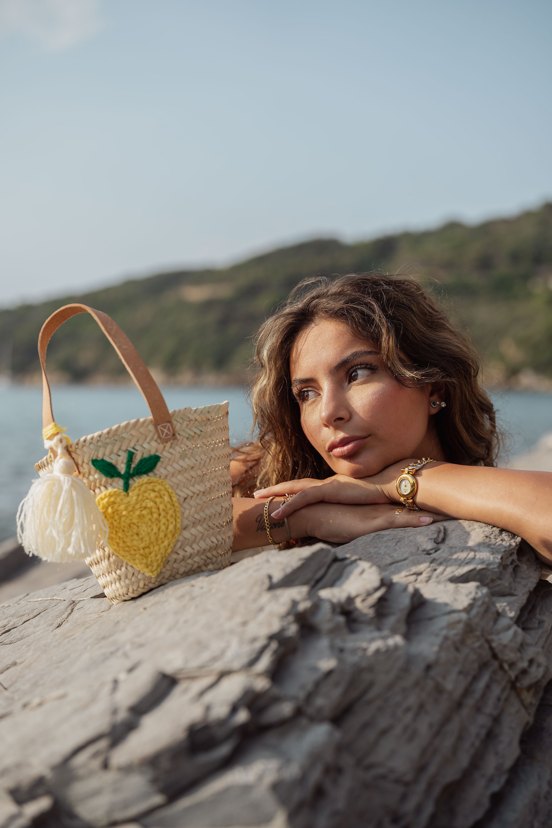 Mini borsa in paglia con ricamo limone giallo appoggiata su roccia vicino al mare con modella in primo piano