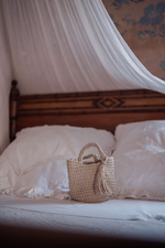 Mini borsa intrecciata a mano Marika Selection modello Sorrento beige con nappa tono su tono appoggiata su letto, accessorio chic artigianale.