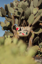Borsa in paglia J’Adore con ricami artigianali, doppia nappa panna e nera appesa a un cactus, stile bohémien mediterraneo.