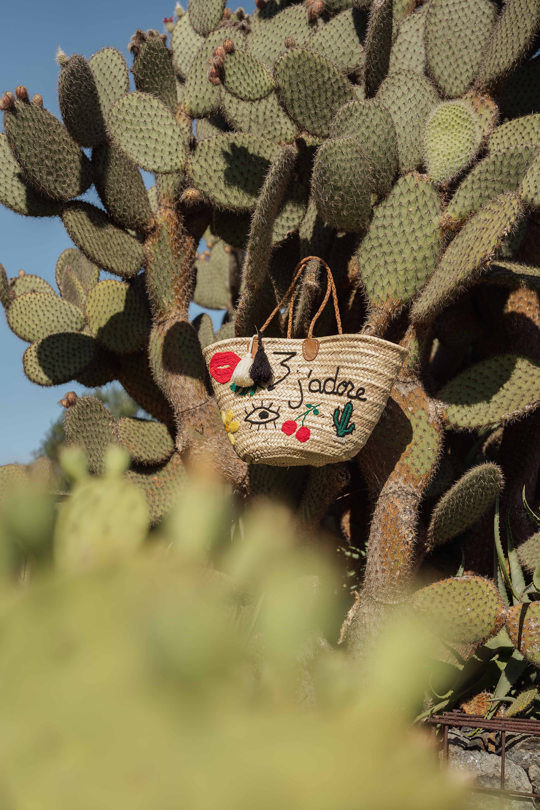 Borsa in paglia J’Adore con ricami artigianali, doppia nappa panna e nera appesa a un cactus, stile bohémien mediterraneo.