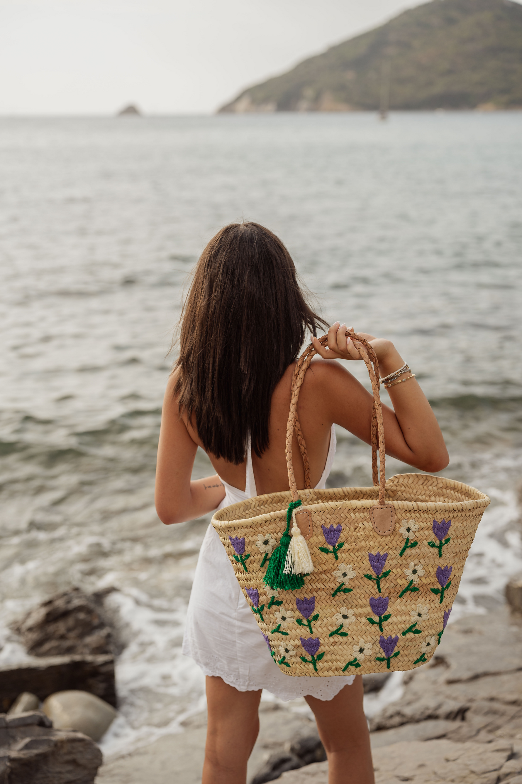 Donna di spalle con borsa in paglia Marika Selection ricamata con fiori viola e bianchi, nappa decorativa verde, vista mare, stile estivo elegante.