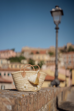 Mini borsa in paglia Marika Selection con ricamo margherite avorio e doppia nappa decorativa avorio e oro appoggiata sulle mura della città.