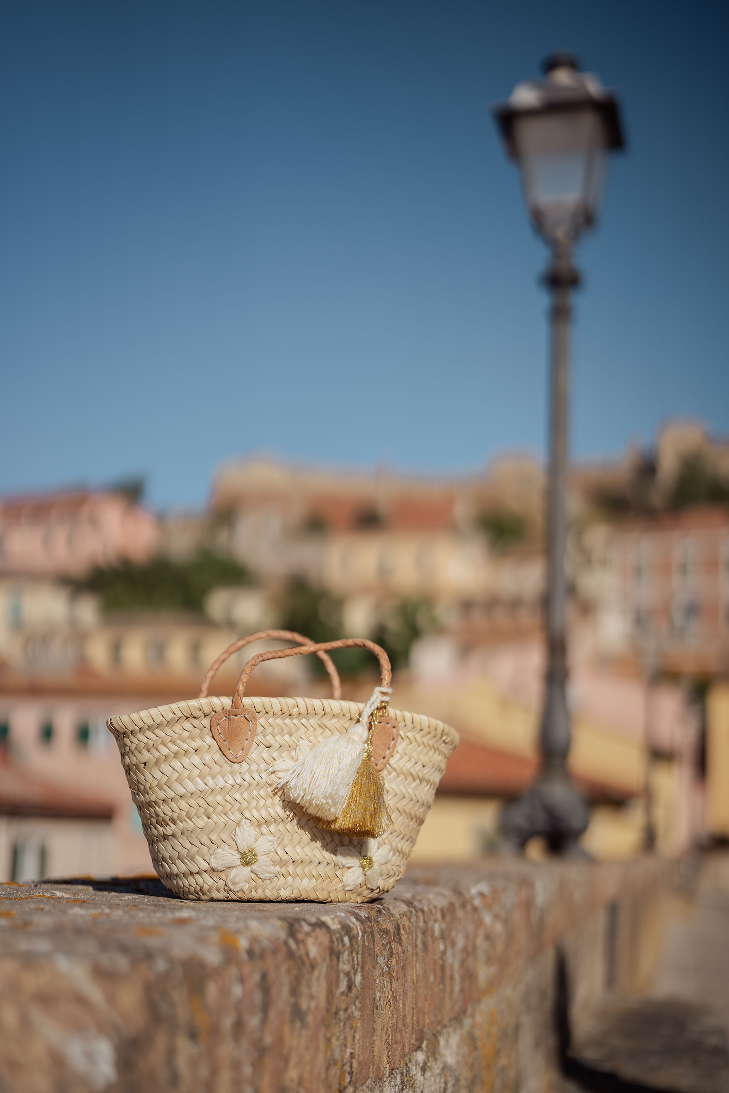 Mini borsa in paglia Marika Selection con ricamo margherite avorio e doppia nappa decorativa avorio e oro appoggiata sulle mura della città.