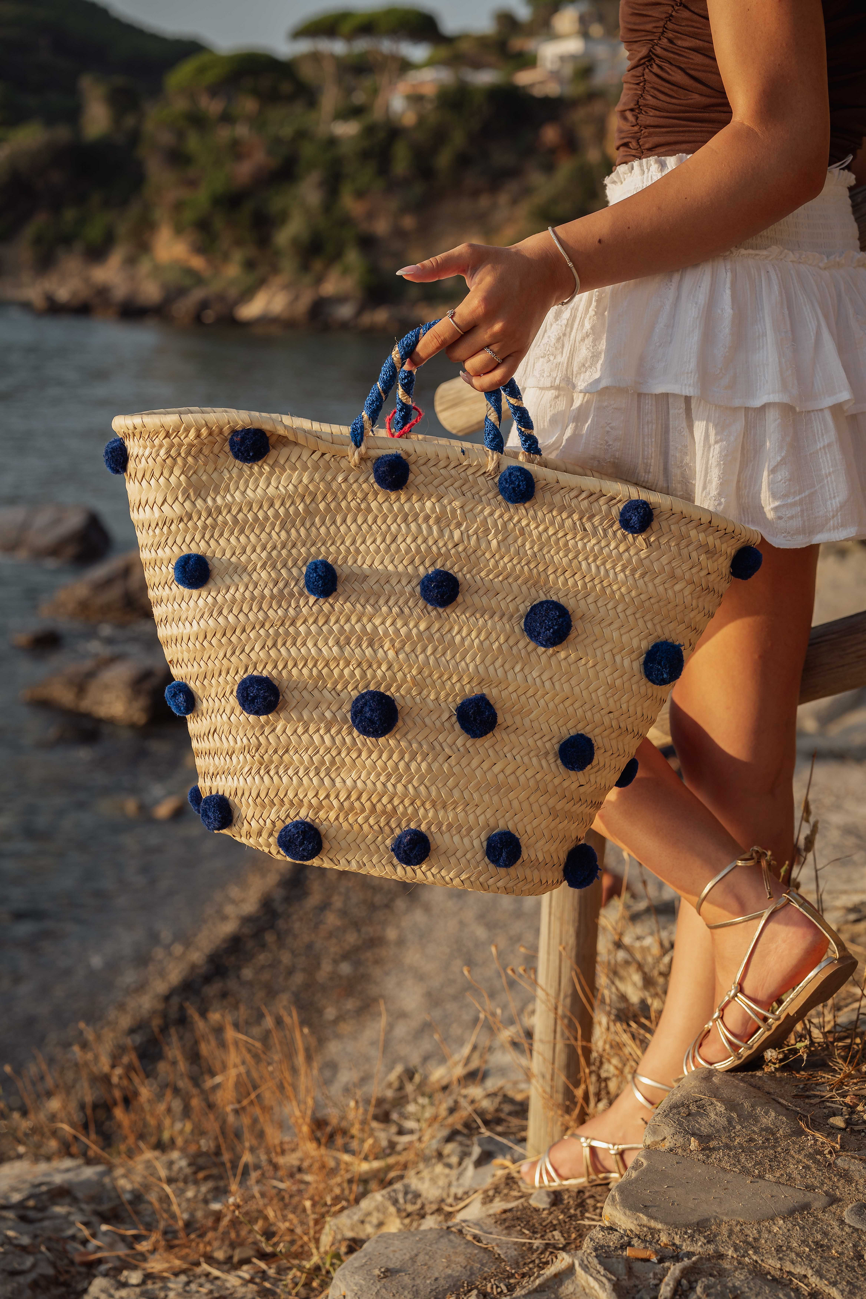 Donna con borsa in paglia decorata con pon pon blu, vista mare su scogliera, stile estivo mediterraneo.
