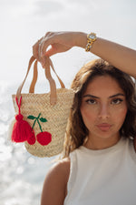 Modella che tiene una mini borsa in paglia con ricamo di ciliegie rosse e nappina rossa davanti al mare al tramonto