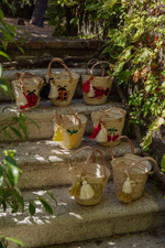 Mini borse in paglia artigianali disposte su scalinata in pietra con ricami limone, ciliegie, coccinella, aragosta, margherite e conchiglia immerse nel verde