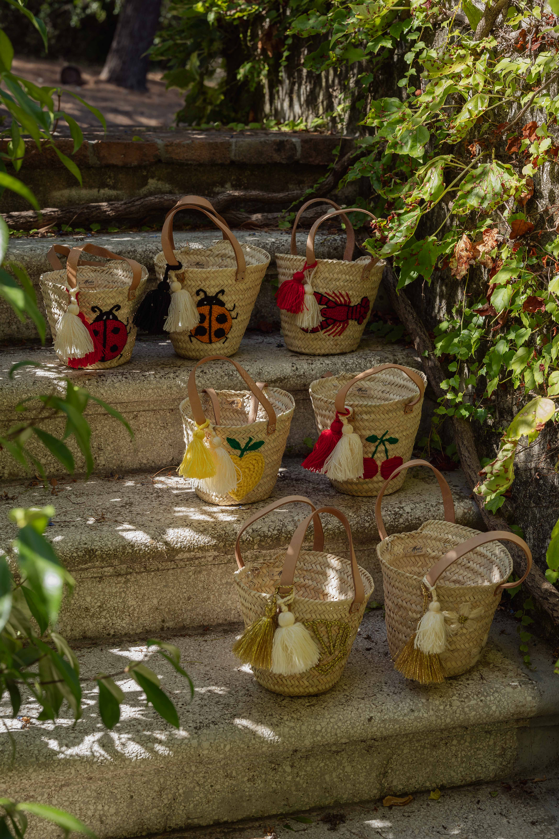 Mini borse in paglia artigianali disposte su scalinata in pietra con ricami limone, ciliegie, coccinella, aragosta, margherite e conchiglia immerse nel verde