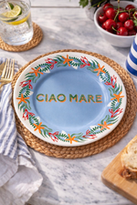Piatto in porcellana Ø16 cm con scritta “Ciao Mare” e decoro marino con stelle marine e coralli, apparecchiato su tavola in marmo con tovaglietta intrecciata, limonata e ciliegie.