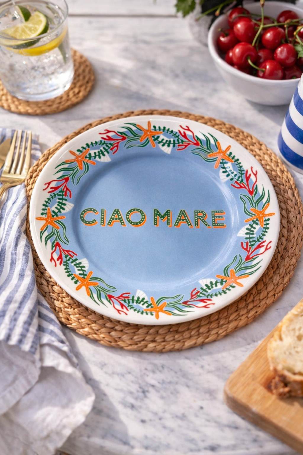 Piatto in porcellana Ø16 cm con scritta “Ciao Mare” e decoro marino con stelle marine e coralli, apparecchiato su tavola in marmo con tovaglietta intrecciata, limonata e ciliegie.
