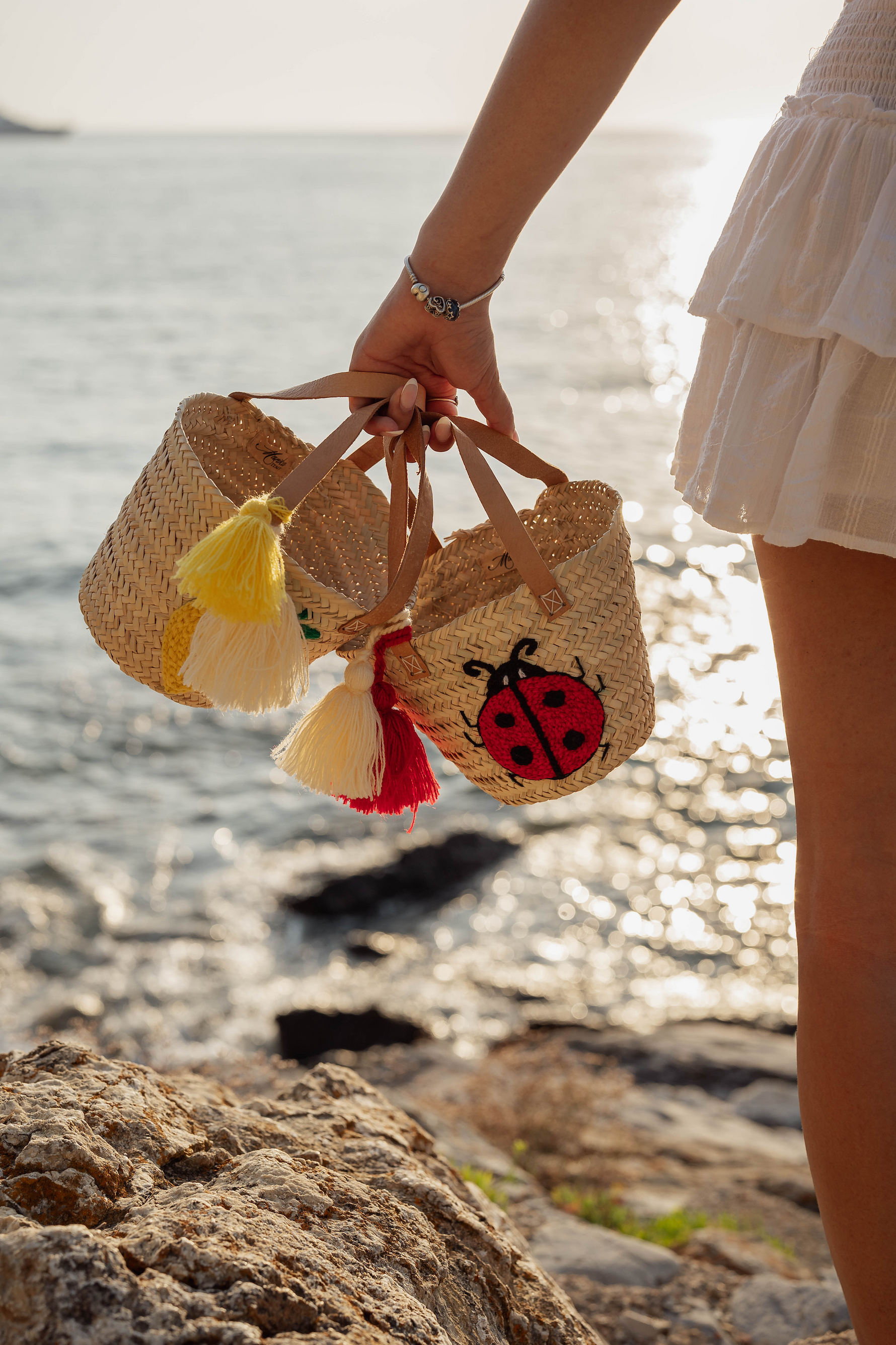 Ragazza che tiene due mini borse in paglia con ricami limone giallo e coccinella rossa davanti al mare al tramonto