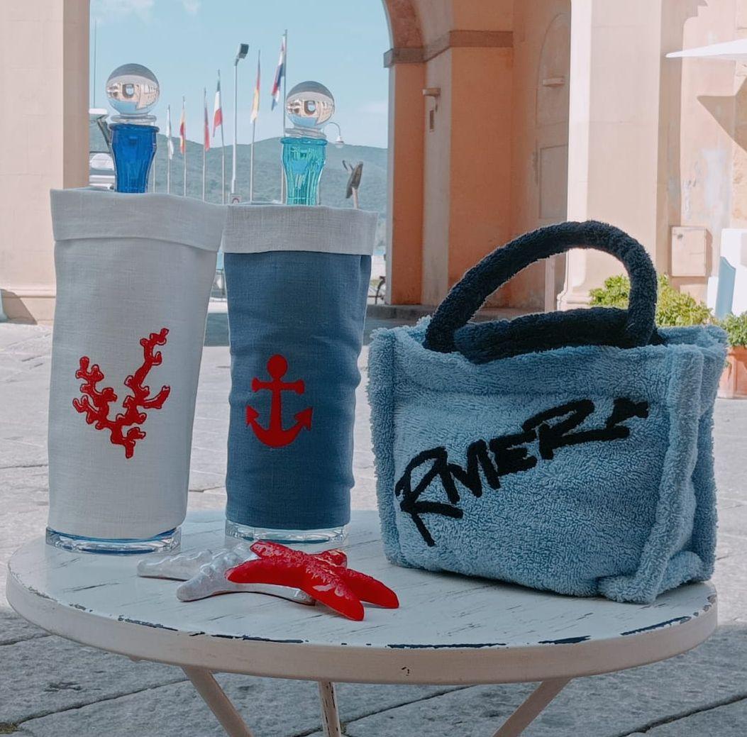 copri bottiglia in lino cerato bianco e blu con ricamo corallo e ancora, borsa in spugna e stella marina decorativa su tavolino estivo

