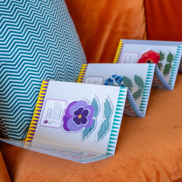 Confezioni Funny Flowers di Vesta aperte, contenenti fiori decorativi in acrilico colorato, appoggiate su un divano arancione accanto a un cuscino a zigzag.