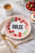 Piatto dessert in porcellana Ø16 cm con scritta “Dolce” e motivo pomodori , ambientato su tavola in marmo con tazza di caffè, fragole e cucchiaino dorato.