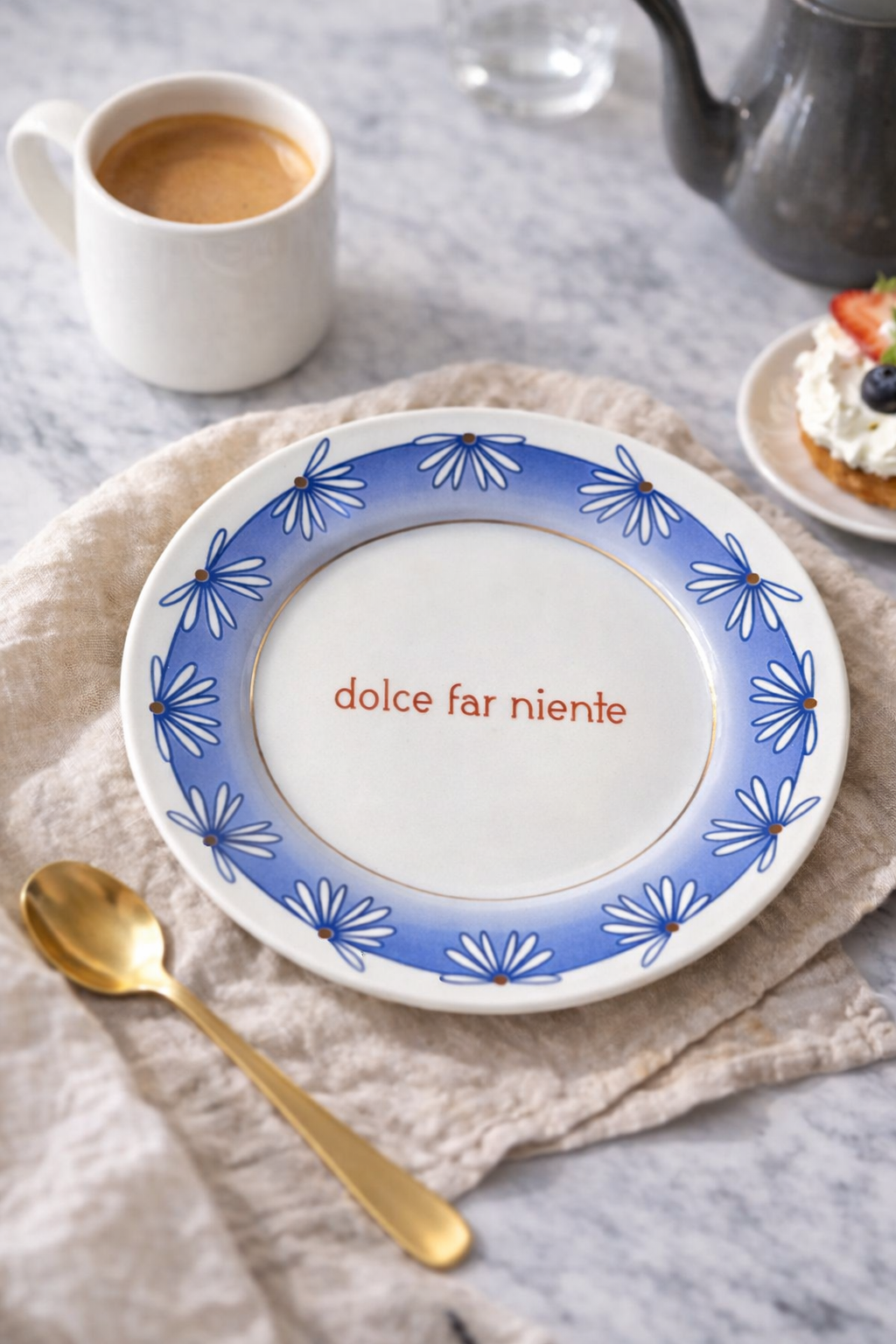 Piatto in porcellana Ø21 cm con scritta “dolce far niente” e bordo azzurro decorato con fiori blu, apparecchiato su tavolo in marmo con tazza di caffè e cucchiaino dorato.